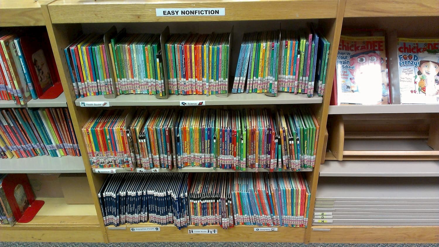 A light oak wooden library bookshelf with 3 metal shelves filled with short books. A large white label on top of the shelf reads "EASY NONFICTION" and more labels on the edge of the metal shelves read Health Books, Science, Holidays, Animals, Biographies (People), Career Books, and Geography.