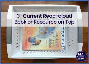 Indigo text overlay reading "3. Current Read-aloud Book or Resource on Top" over a photo of a white plastic storage bin with a library books and printed lesson materials in it, sitting on a dark wood background