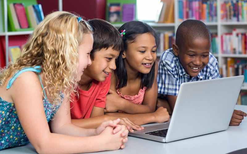 Four children with diverse skin colors using a laptop in the school library.